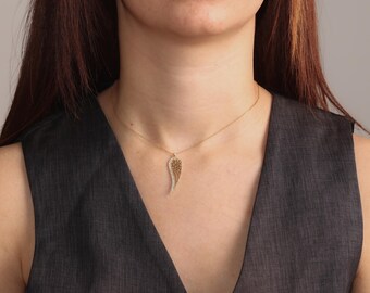 May include: A gold-colored necklace with a pendant shaped like a wing. The wing pendant is detailed with small, sparkling crystals along one edge and intricate patterns. The necklace is worn against a dark gray top.