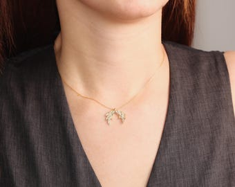 May include: A delicate gold-colored necklace featuring a pendant shaped like two stylized leaves or wings. The pendant is adorned with small, sparkling accents. The necklace is displayed against a dark gray background, highlighting the jewelry's design.
