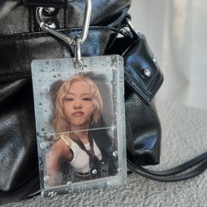 May include: Black leather handbag with silver hardware and a clear acrylic tag. The tag features a photo of a person with blonde hair. The bag has a top handle and a shoulder strap.