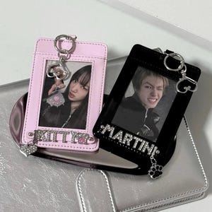 May include: A silver-colored, faux leather wallet with a magnetic clasp. Two ID card holders, one pink and one black, are on top. The pink holder has the word "KITTY" and a heart charm. The black holder has the word "MARTINI" and a paw charm.
