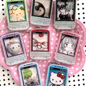 Puede incluir: Una colección de llaveros en miniatura con forma de teléfono y colores vibrantes. Cada llavero presenta un diseño diferente dentro de la pantalla, incluyendo personajes de dibujos animados e ilustraciones. Los llaveros tienen un diseño de teclado QWERTY y están unidos a una cadena plateada.