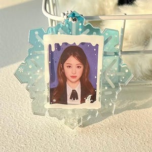 Pode incluir: Um porta-chaves transparente em forma de floco de neve com uma foto de uma pessoa no interior. O floco de neve é azul claro com detalhes brancos e uma corrente prateada. A foto mostra uma pessoa com cabelo castanho usando um blazer preto e uma camisa branca.