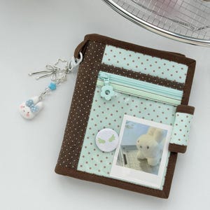 May include: A bifold wallet featuring a light blue and brown polka dot pattern, a clear front pocket with a zipper, a flower-shaped zipper pull, a circular pin with wing graphics, and a photo of a white plush rabbit tucked into a clear window.