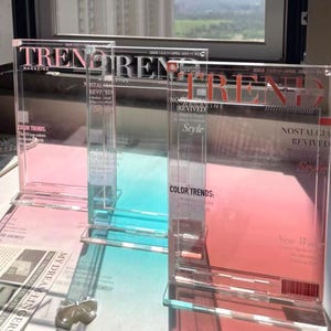 Könnte beinhalten: Transparente Acryl-Zeitschriftenhalter mit dem Wort "TREND" in Rot. Die Zeitschriften haben ein Farbdesign in Rosa, Blau und Weiß. Der Text "COLOR TRENDS" und "NOSTALGIA REVIVED" sind ebenfalls sichtbar.