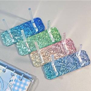May include: A collection of colorful acrylic display stands in shades of blue, green, pink, and yellow. Each stand features a textured surface and a clear, upright support for holding items. The stands are designed to showcase small objects.