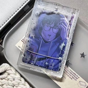May include: A clear, rectangular keychain featuring a manga-style illustration of a person with glasses and a blue hoodie. The image is encased in a clear, textured plastic. The keychain is on a silver tray with a white tag and a silver case.