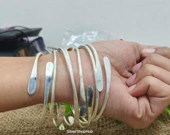 Puede incluir: Un conjunto de pulseras de metal plateado con un diseño único. Cada pulsera tiene un diseño curvo y abierto con un extremo plano y alargado. Las pulseras están apiladas en una muñeca, mostrando su superficie reflectante.