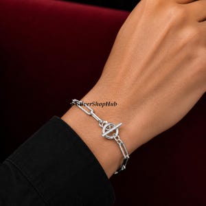Op de afbeelding: Een zilverkleurige kettingarmband met langwerpige, rechthoekige schakels en een ronde knevelsluiting. Het metalen oppervlak heeft een gepolijste afwerking en de tekst SilverShopHub is zichtbaar bij de sluiting op de schakels van de armband.