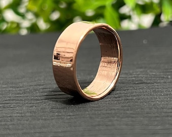 May include: A rose gold ring with a flat band. The ring is made of a shiny metal and has a smooth, polished surface. The ring is set against a dark background with green foliage in the background.