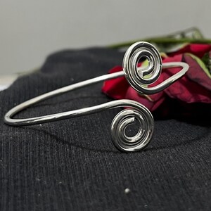 May include: A silver cuff bracelet featuring a spiral design at each end. The open bracelet is curved and set against a dark background with a red rose.