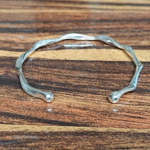 Puede incluir: Un brazalete abierto de color plateado con una textura ondulada y orgánica. La banda de metal presenta un acabado pulido y reflectante con extremos redondeados y suaves. El diseño es circular con una pequeña abertura en un extremo.