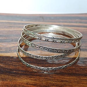 Może przedstawiać: Zestaw czterech ułożonych w stos bransoletek typu bangle ze srebrzystego metalu. Każda bransoletka ma inny wytłoczony wzór, w tym motywy kwiatowe, powtarzające się kształty geometryczne i małe figurki delfinów, z polerowanym wykończeniem.