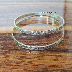 May include: Two silver-toned metal bangles featuring intricate embossed patterns. The top bangle displays a repeating circular motif, while the bottom bangle showcases an elongated floral design. The text SilverShopHub is visible in the upper right.
