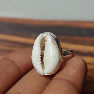 May include: A ring featuring a white cowrie shell set in a polished silver-toned metal band. The shell is oval-shaped with a central longitudinal slit and serrated edges, securely mounted within a smooth metal bezel setting on a simple band.