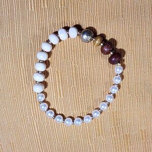 Puede incluir: Una pulsera de cuentas blancas, perlas y marrones. La pulsera incluye una cuenta plateada con un diseño de hojas. Las cuentas están ensartadas en un cordón elástico, creando un accesorio flexible y cómodo.