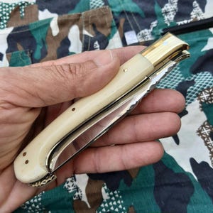 Könnte beinhalten: Ein Klappmesser mit cremefarbenem Griff und glänzender, silberner Klinge. Das Messer hat eine goldfarbene Zwinge und Nieten. Die Klinge ist teilweise geöffnet und zeigt ihre scharfe Schneide. Das Messer wird in einer Hand vor einem Tarnhintergrund gehalten.
