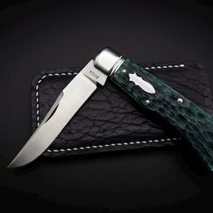 May include: A folding pocket knife with a single silver-toned metal blade featuring the word RITCHIE engraved near the bolster. The handle has a textured dark green finish with a silver-toned shield inlay and polished metal bolsters at the end.