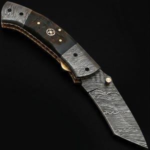 May include: A folding pocket knife featuring a blade and bolster with a distinct wavy Damascus steel pattern. The handle is constructed from dark wood with brass pins and a central circular mosaic inlay. A small brass thumb stud is attached to the blade.