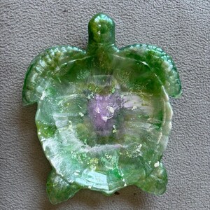 Puede incluir: Un plato decorativo con forma de tortuga. El plato está hecho de resina verde translúcida con detalles blancos y morados. La tortuga tiene una superficie texturizada y está diseñada para guardar objetos pequeños.