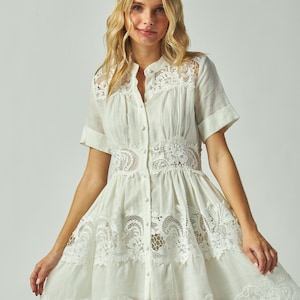 May include: A white, short-sleeved dress with intricate lace detailing. The dress features a button-down front, a high collar, and lace accents around the waist and hem. The dress is made of a lightweight fabric.