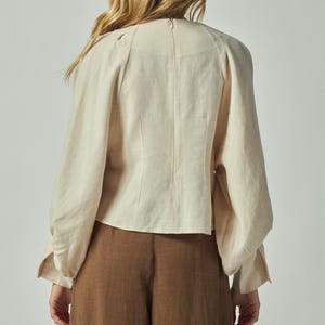May include: Back view of a cream-colored blouse with long sleeves and a high neckline. The blouse has a fitted bodice and voluminous sleeves. Paired with brown wide-leg pants, the outfit is simple and elegant.