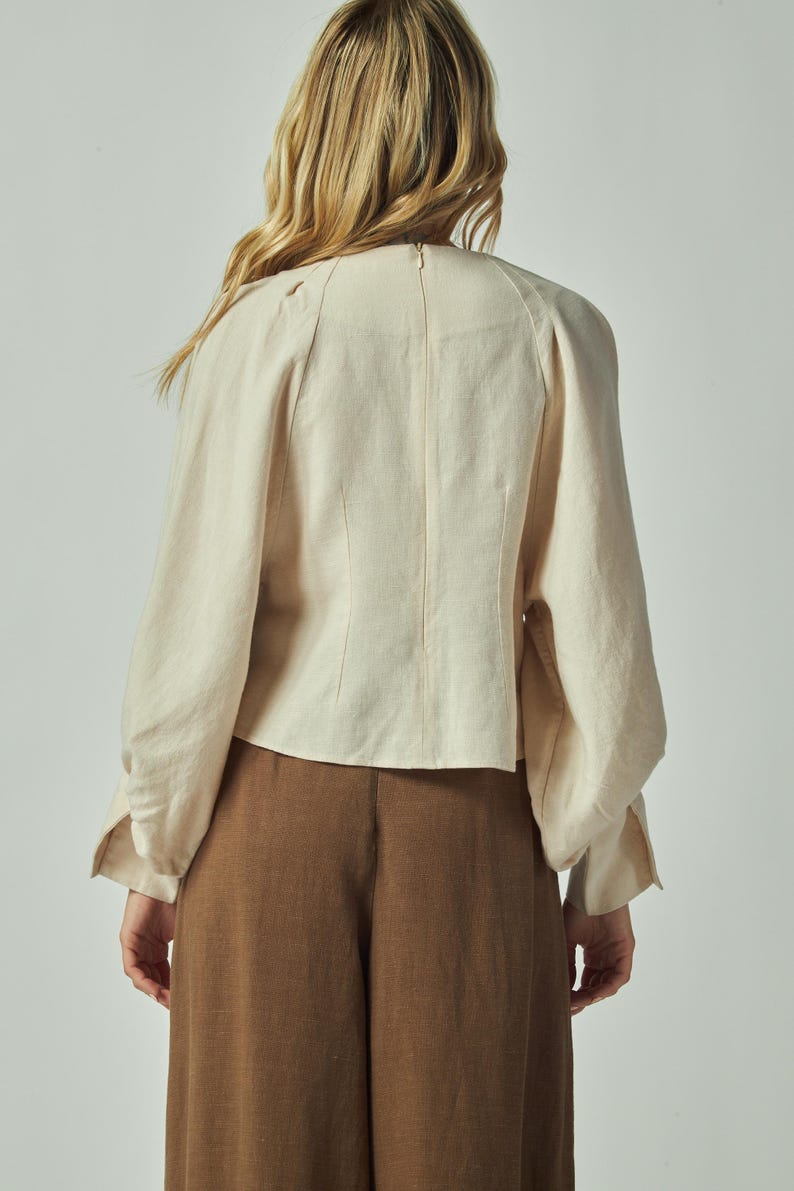 May include: Back view of a cream-colored blouse with long sleeves and a high neckline. The blouse has a fitted bodice and voluminous sleeves. Paired with brown wide-leg pants, the outfit is simple and elegant.
