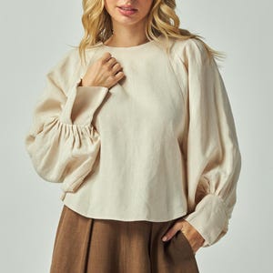 May include: Cream-colored linen blouse with long sleeves and a round neckline. The sleeves have a gathered detail at the elbow and a buttoned cuff. The blouse is paired with brown pants. Fashion apparel.