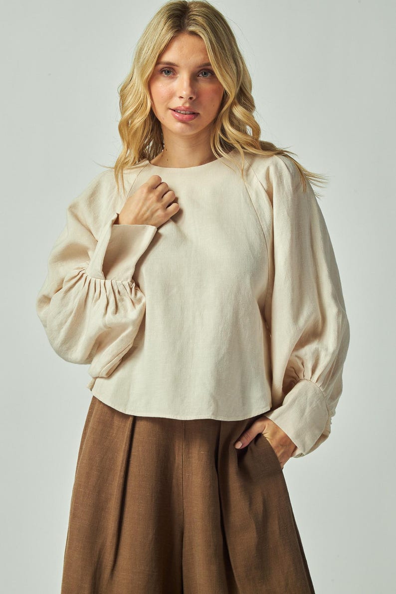 May include: Cream-colored linen blouse with long sleeves and a round neckline. The sleeves have a gathered detail at the elbow and a buttoned cuff. The blouse is paired with brown pants. Fashion apparel.