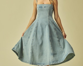 May include: Light blue denim dress with a square neckline and a flared skirt. The dress features thin straps and visible seams. The model is wearing brown sandals. The dress is a classic, timeless piece.