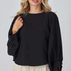 May include: Black long-sleeve top with a round neckline and gathered sleeves. The top is paired with light-colored pants. The sleeves have a cuff at the wrist. The top is made of a soft, comfortable material.