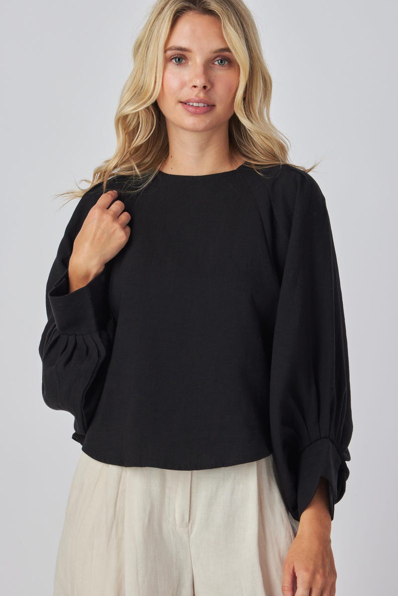 May include: Black long-sleeve top with a round neckline and gathered sleeves. The top is paired with light-colored pants. The sleeves have a cuff at the wrist. The top is made of a soft, comfortable material.