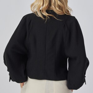 May include: Black long-sleeved top with a rounded neckline and gathered sleeves. The top is made of a textured fabric and has a tailored fit. The model is wearing light-colored pants.