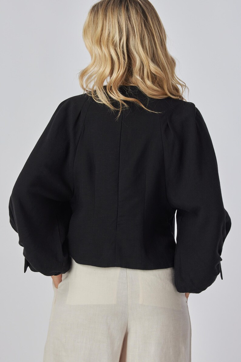 May include: Black long-sleeved top with a rounded neckline and gathered sleeves. The top is made of a textured fabric and has a tailored fit. The model is wearing light-colored pants.