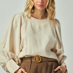 May include: A beige long-sleeve blouse with a round neckline and voluminous sleeves. The blouse is paired with brown high-waisted pants and a woven belt with a wooden buckle. The outfit is a neutral color palette.