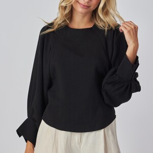 May include: Black long-sleeve top with a round neckline and gathered sleeves. The top is paired with light-colored pants. The sleeves have a cuff detail. The top is made of a solid black fabric.
