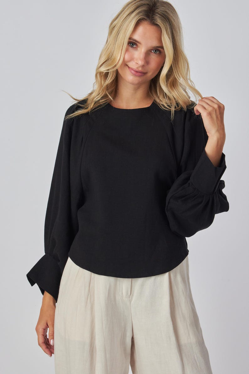 May include: Black long-sleeve top with a round neckline and gathered sleeves. The top is paired with light-colored pants. The sleeves have a cuff detail. The top is made of a solid black fabric.
