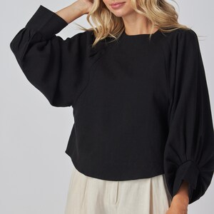 May include: A black long-sleeved top with a round neckline and voluminous sleeves. The top is paired with light-colored pants. The sleeves have a gathered detail at the wrist. The model is wearing the top.