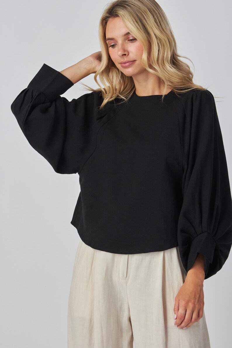 May include: A black long-sleeved top with a round neckline and voluminous sleeves. The top is paired with light-colored pants. The sleeves have a gathered detail at the wrist. The model is wearing the top.