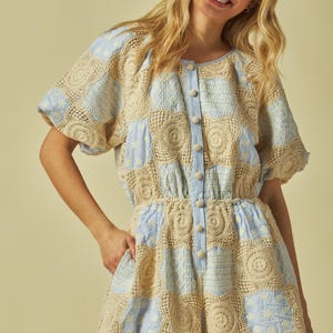 May include: A light blue and beige patchwork romper with a button-up front. The romper features a combination of crochet and patterned fabric, with short sleeves and shorts. The garment has a fitted waist and pockets.
