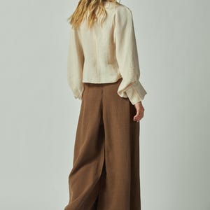 May include: A woman wearing a cream-colored blouse with long sleeves and brown wide-leg pants. The blouse has a button-down front and the pants have a high waist. The outfit is made of a soft, flowing fabric.