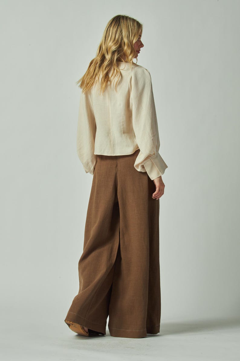 May include: A woman wearing a cream-colored blouse with long sleeves and brown wide-leg pants. The blouse has a button-down front and the pants have a high waist. The outfit is made of a soft, flowing fabric.