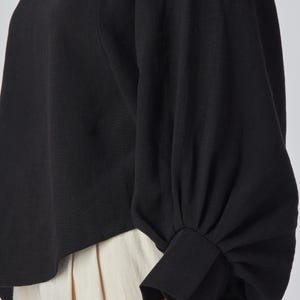 May include: Black long-sleeved top with a round neckline and a gathered sleeve design. The sleeves have a cuff detail at the wrist. The bottom of the top is visible, paired with cream-colored pants.