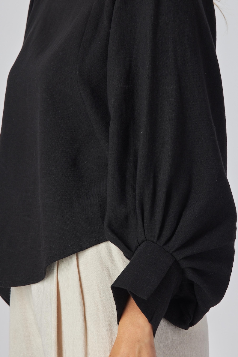 May include: Black long-sleeved top with a round neckline and a gathered sleeve design. The sleeves have a cuff detail at the wrist. The bottom of the top is visible, paired with cream-colored pants.