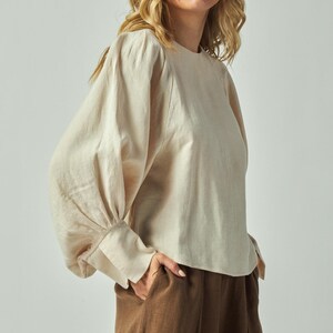 May include: A beige long-sleeved blouse with a round neckline and gathered sleeves. The blouse is paired with brown pleated pants. The model is wearing the outfit in a studio setting.
