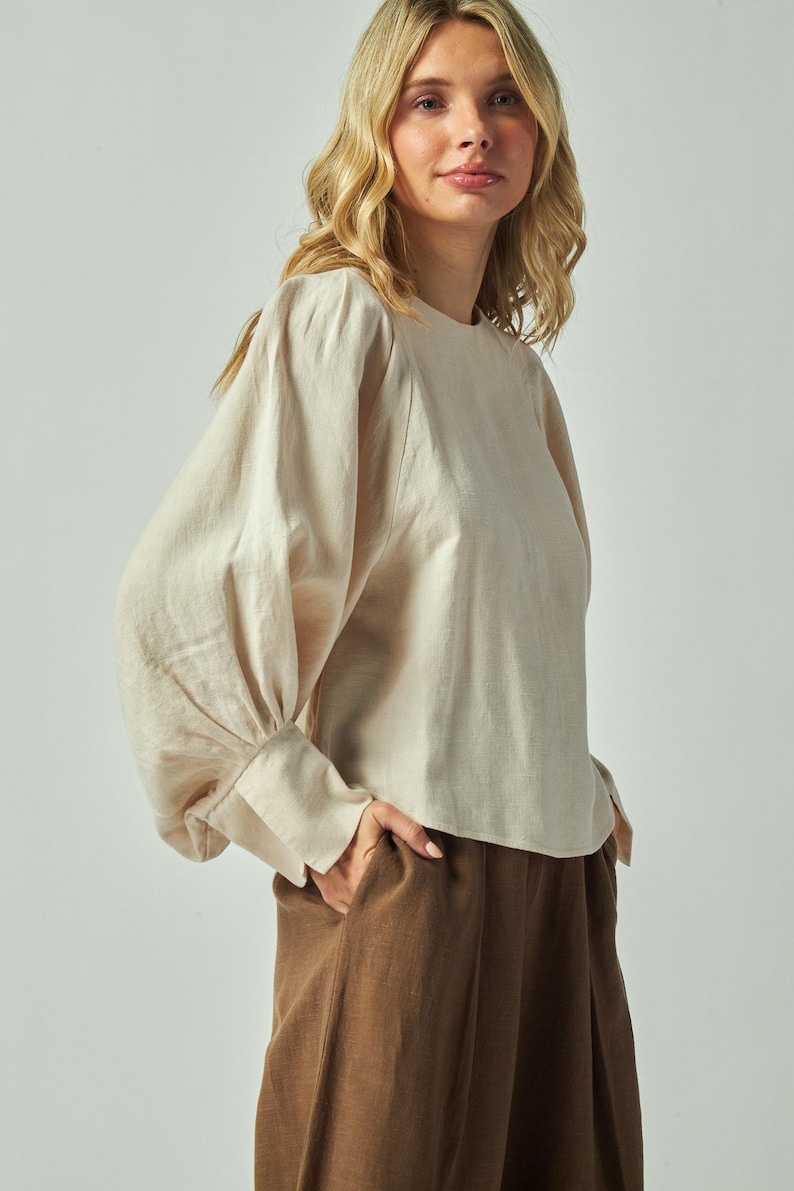 May include: A beige long-sleeved blouse with a round neckline and gathered sleeves. The blouse is paired with brown pleated pants. The model is wearing the outfit in a studio setting.