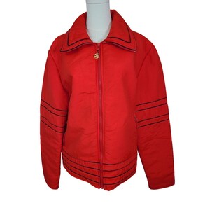 May include: A bright red jacket with a zip-up front and a wide collar. The jacket features black stitched detailing on the sleeves and around the waist. A gold-colored emblem is on the zipper pull.