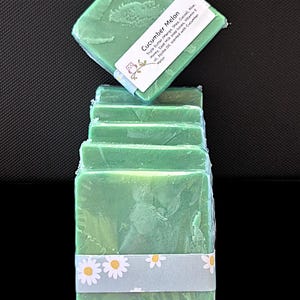 May include: A stack of green, square soaps, each wrapped in clear plastic and secured with a band decorated with daisies. The top soap has a label reading "Cucumber Melon". The soaps are a vivid green colour.