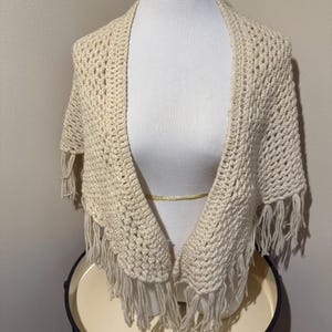 May include: Cream-colored, open-weave crochet shawl with fringe. The shawl is draped over a mannequin, showcasing its open front design and textured pattern. The fringe hangs along the bottom edge, adding a decorative element. This handmade shawl is a cozy accessory.