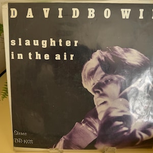 May include: A vintage vinyl record album cover for David Bowie, titled "slaughter in the air." The cover is black with white text and a grayscale image of Bowie. The record is in a plastic sleeve.