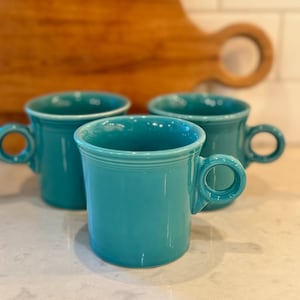 May include: Three turquoise ceramic mugs with rounded handles are arranged on a light-colored surface. The mugs have a classic design with a ring detail near the rim. A wooden cutting board is in the background.